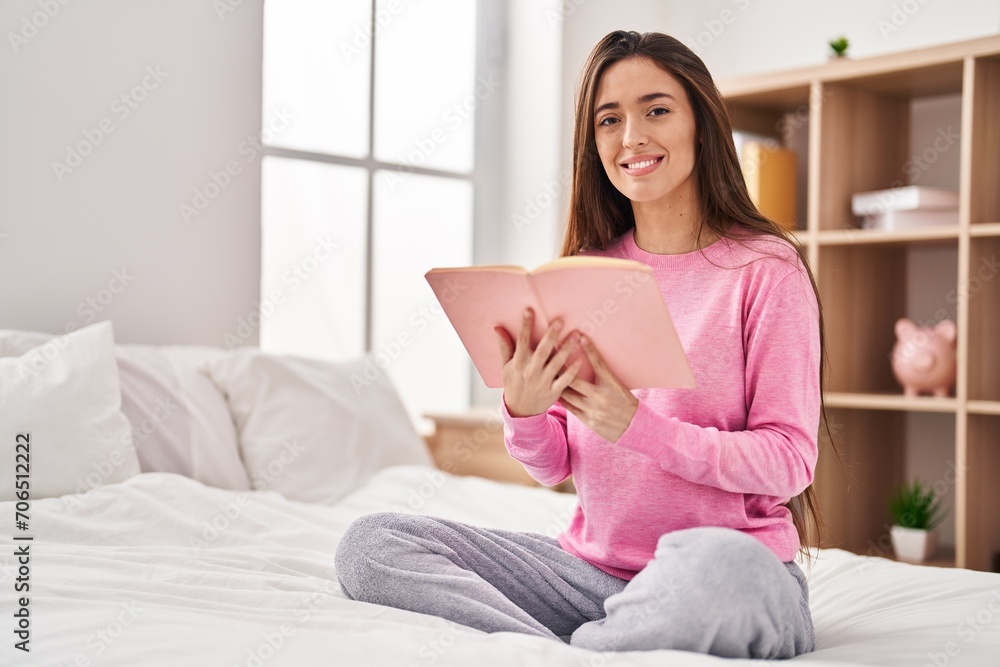 Young beautiful hispanic woman reading book sitting on bed at bedroom