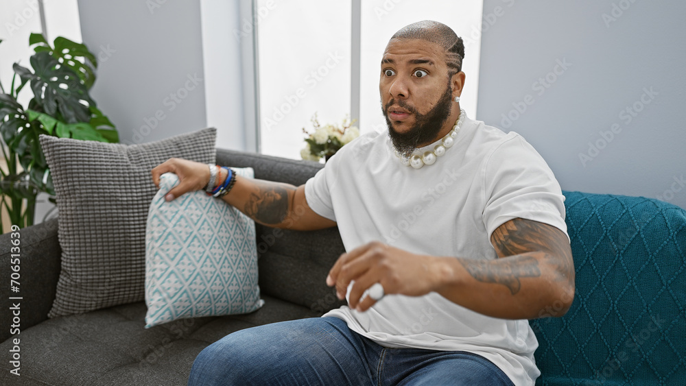 A surprised african american man with a beard sits in a living room, portraying a relaxed yet expressive moment. A surprised african american man with a beard sits in a living room, portraying a relaxed yet expressive moment.