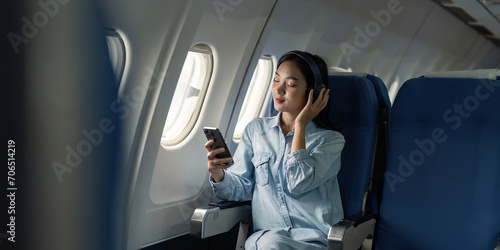Asian woman traveler in airplane wearing headset listening music from mobile phone going on a trip vacation travel concept