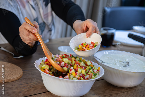 The wife fills a salad bowl with a large bowl of yogurt next to it to provide the guests with what they need at lunchtime at the large collective table for the large family.