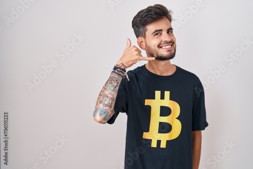 Young hispanic man with tattoos wearing bitcoin t shirt smiling doing phone gesture with hand and fingers like talking on the telephone. communicating concepts.