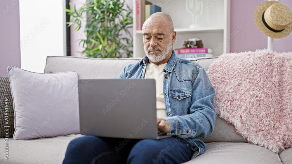 A middle-aged man with grey beard in denim jacket using laptop on sofa at cozy home interior A middle-aged man with grey beard in denim jacket using laptop on sofa at cozy home interior