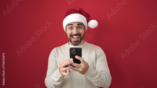 Young hispanic man wearing christmas hat using smartphone over isolated red background © Krakenimages.com
