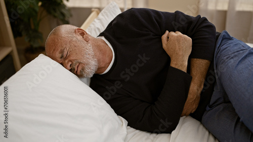 A mature hispanic man with gray hair and a beard clutches his abdomen in pain while lying in a bedroom. photo