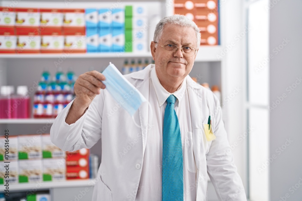 Middle age grey-haired man pharmacist smiling confident holding medical mask at pharmacy
