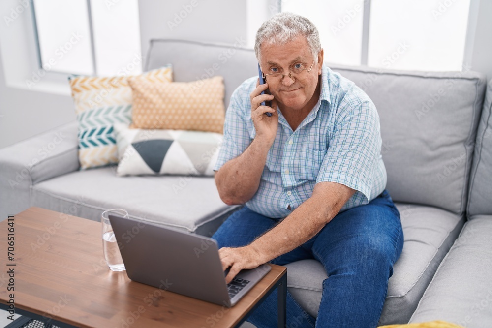 Middle age grey-haired man talking on smartphone using laptop sitting on sofa at home Middle age grey-haired man talking on smartphone using laptop sitting on sofa at home