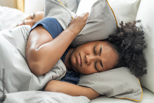 black African American woman covers her ears with a pillow, can't sleep, Asian husband's snoring sounds loudly disturbing her sleep on the bed at home. photo