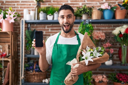 Hispanic young man working at florist shop showing smartphone screen afraid and shocked with surprise and amazed expression, fear and excited face.