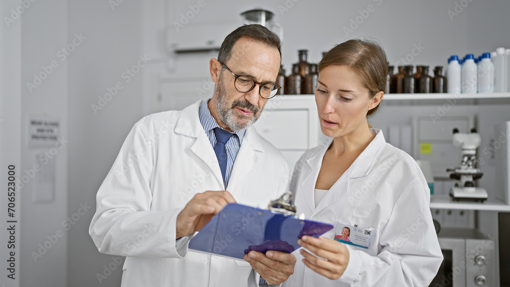 Serious chemists in lab, two science partners engrossed in reading crucial document on clipboard, immersed in research work together in indoor laboratory Serious chemists in lab, two science partners engrossed in reading crucial document on clipboard, immersed in research work together in indoor laboratory