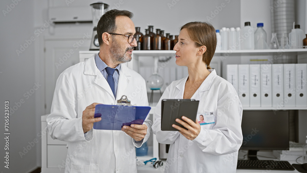 Two serious scientists in the lab, reading experiment document on clipboard, speaking and collaborating using touchpad in laboratory workplace. Two serious scientists in the lab, reading experiment document on clipboard, speaking and collaborating using touchpad in laboratory workplace.