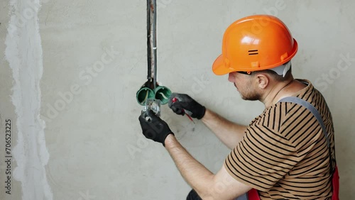 Electrician male in uniform, protective gloves and helmet checks presence of electrical voltage in socket phase uses electrical tester screwdriver. Examining wires in outlet by voltage detector indoor photo
