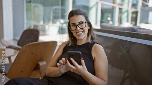 Radiant hispanic woman, all smiles, busily messaging on her smartphone while fashionably lounging in a modern hotel lobby