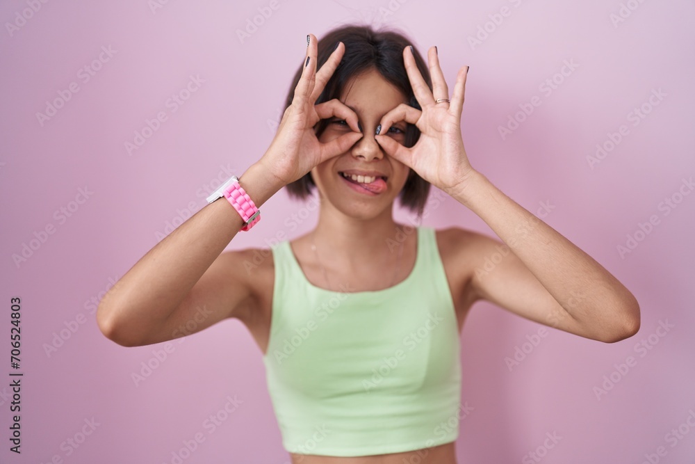 Young girl standing over pink background doing ok gesture like binoculars sticking tongue out, eyes looking through fingers. crazy expression. Young girl standing over pink background doing ok gesture like binoculars sticking tongue out, eyes looking through fingers. crazy expression.
