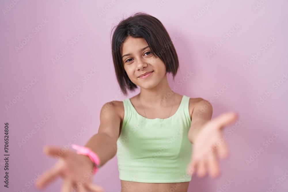 Young girl standing over pink background smiling cheerful offering hands giving assistance and acceptance.