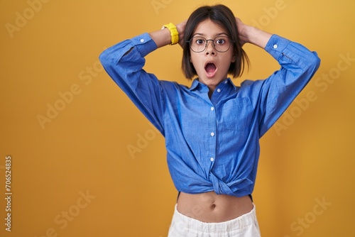 Young girl standing over yellow background crazy and scared with hands on head, afraid and surprised of shock with open mouth photo