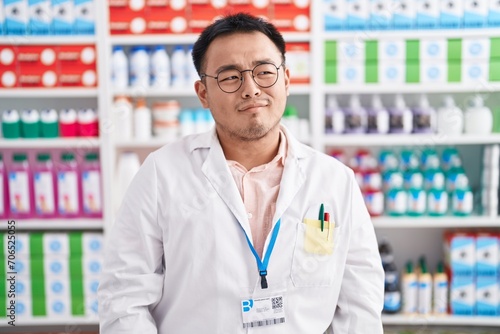 Chinese young man working at pharmacy drugstore smiling looking to the side and staring away thinking. © Krakenimages.com