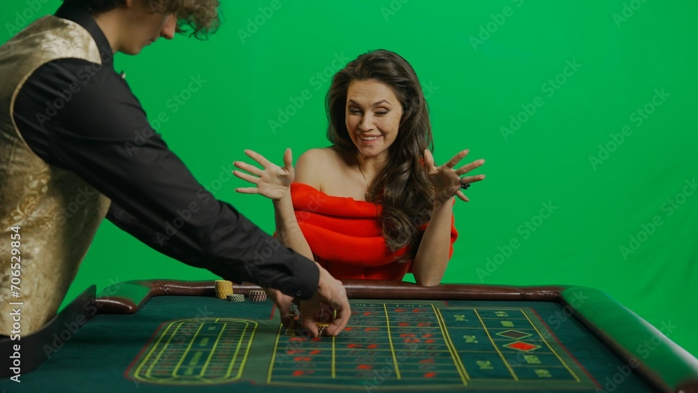 Gorgeous female in studio on chroma key green screen. Appealing woman in red dress and male croupier at the roulette table takes winning money. Gorgeous female in studio on chroma key green screen. Appealing woman in red dress and male croupier at the roulette table takes winning money.