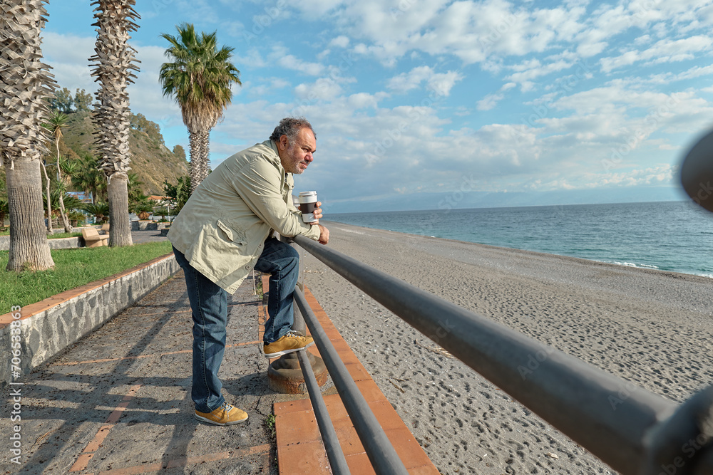 Pensive middle-aged bearded man drinking coffee on seaside in sunny winter or spring day. Pensive middle-aged bearded man drinking coffee on seaside in sunny winter or spring day.