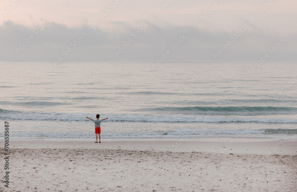 Obraz premium Kleiner Junge mit roten Hosen und blauem Shirt ist alleine am Strand die Hände ausgestreckt schaut auf das Meer und spielt mit den Wellen