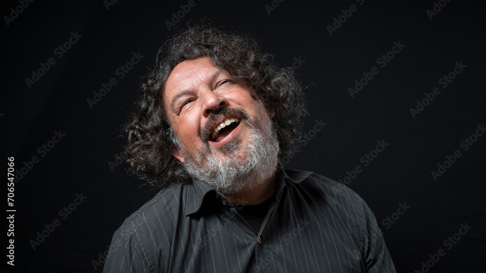 Man with white beard and black curly hair with funny expression, looking up, wearing black shirt against black background