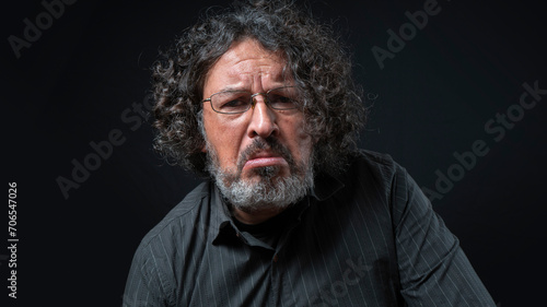 Man with white beard and black curly hair with sad expression, wearing black shirt against black background