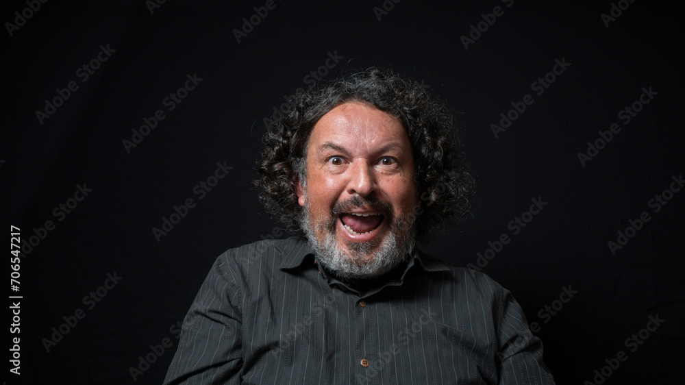 Man with white beard and black curly hair with happy and excited expression, wearing black shirt against black background Man with white beard and black curly hair with happy and excited expression, wearing black shirt against black background