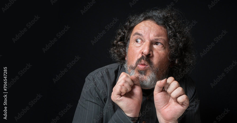 Man with white beard and black curly hair with scared expression, scared looking to the left, wearing black shirt against black background Man with white beard and black curly hair with scared expression, scared looking to the left, wearing black shirt against black background