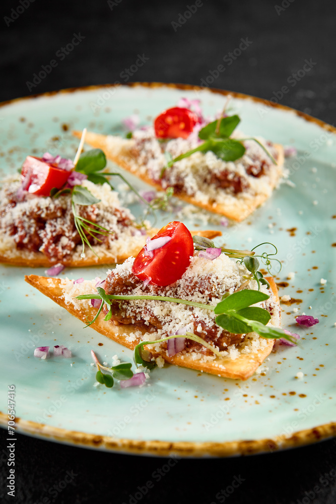 Elegant beef tartare on crispy toasts with parmesan, greens, and tomatoes on a speckled turquoise plate