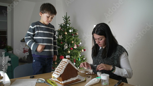 Mother and Child Creating Gingerbread House, Home December Festivities with Christmas Tree in background. Family preparing for holidays