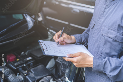 Auto check, car service shop concept. Automobile repairman writing job checklist on clipboard, mechanic checking engine to estimate repair machine, inspecting maintenance by engineer at vehicle garage photo