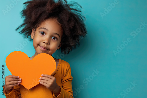 niña afroamericana posando con un corazón de cartulina naranja en sus manos, sobre fondo verde turquesa photo
