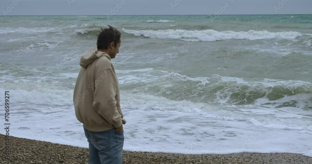 Adult Caucasian male standing on a pebble beach and looking at the stormy sea Adult Caucasian male standing on a pebble beach and looking at the stormy sea