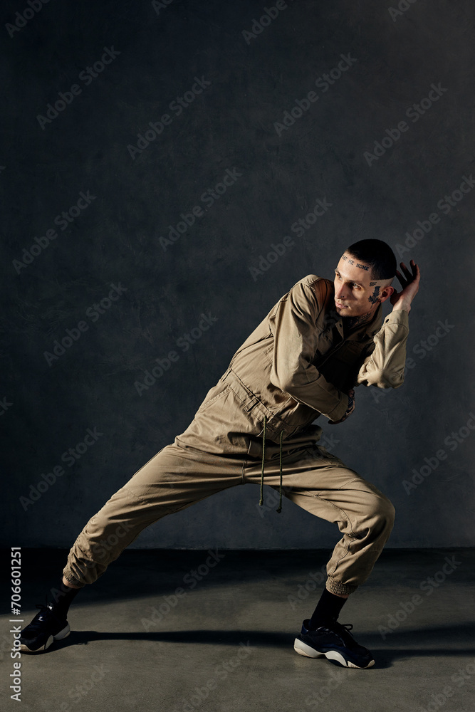 Handsome performer with tattooed body, earrings, beard. Dressed in khaki overalls and black sneakers. Dancing on gray background. Dancehall, hip-hop