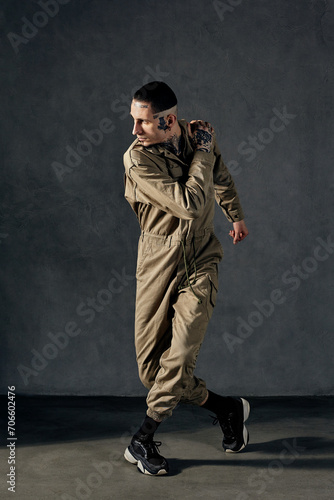 Graceful fellow with tattooed body and face, earrings, beard. Dressed in khaki jumpsuit, black sneakers. Dancing, gray background. Dancehall, hip-hop