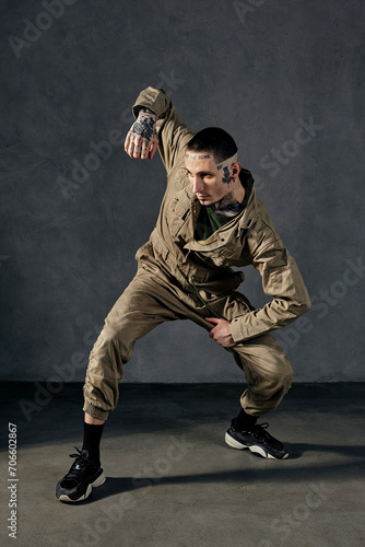 Modern fellow with tattooed body and face, earrings, beard. Dressed in khaki jumpsuit, black sneakers. Dancing on gray background. Dancehall, hip-hop