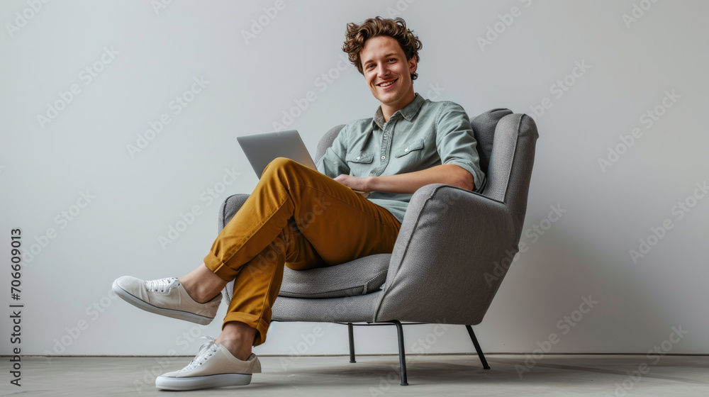Young Caucasian man with curly hair, comfortably seated in a modern gray armchair, smiling and relaxed while using a laptop on his lap - obrazy, fototapety, plakaty 