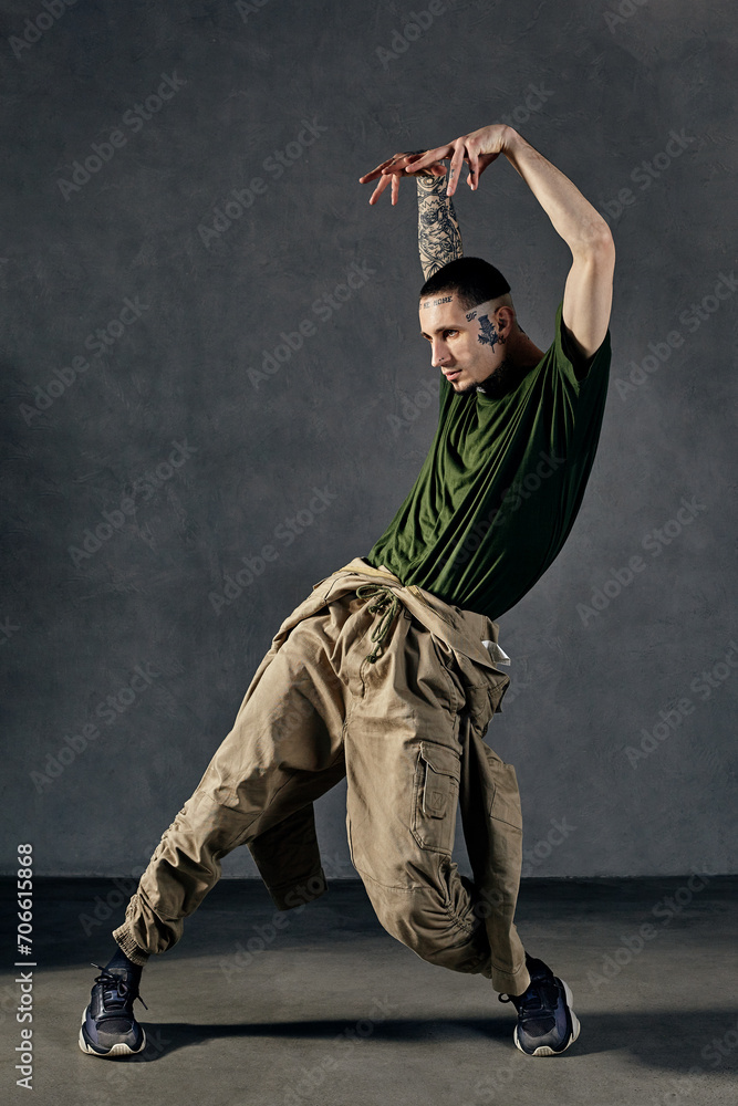 Fellow with tattooed body, earrings, beard. Dressed in khaki t-shirt and jumpsuit, black sneakers. Dancing on gray background. Dancehall, hip-hop