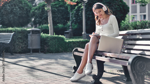 Remote learning. A smiling young woman in summer attire and wireless headphones is taking notes from a lesson displayed on her laptop into her study notebook while seated on a bench in a park