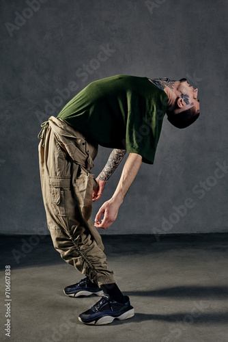 Graceful guy with tattooed body, earrings, beard. Dressed in khaki t-shirt, overalls, black sneakers. Dancing on gray background. Dancehall, hip-hop