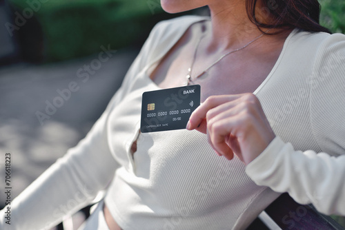 Worldwide banking. A cropped close-up of a beautiful young adult woman in a white outfit holding a black bank credit card, peacefully sitting outdoors on a sunny bench