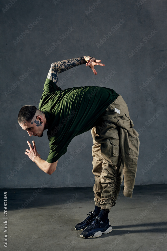 Athletic guy with tattooed body, earrings, beard. Dressed in khaki t-shirt, overalls, black sneakers. Dancing on gray background. Dancehall, hip-hop
