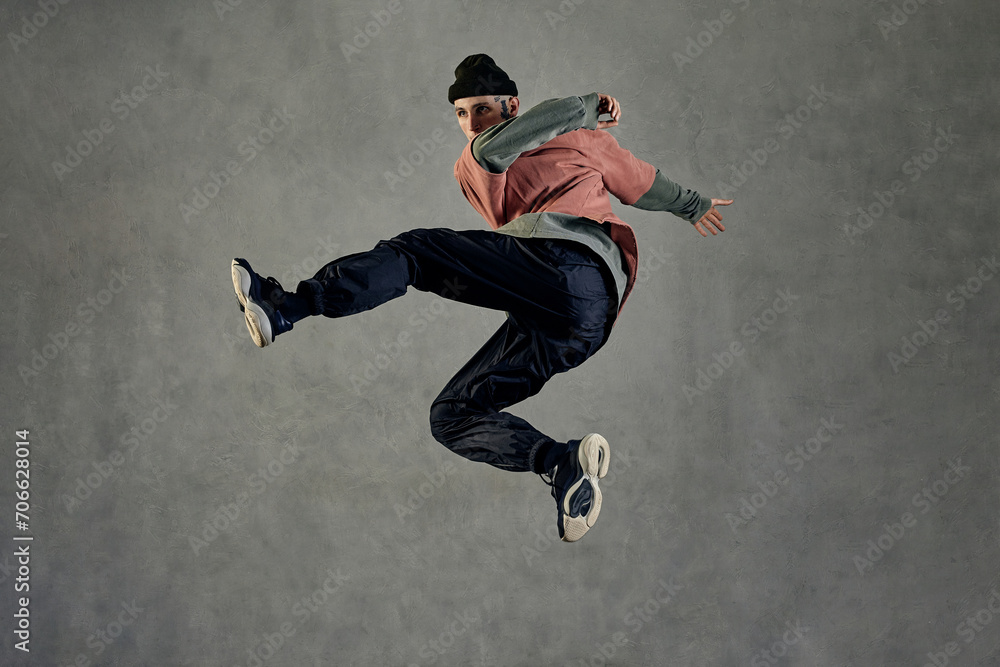 Fellow with tattooed face, beard. Dressed in hat, colorful jumper, black pants and sneakers. Jumping, dancing on gray background. Dancehall, hip-hop
