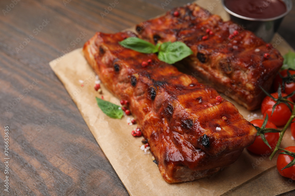 Tasty roasted pork ribs served with sauce, basil and tomatoes on wooden table, closeup. Space for text