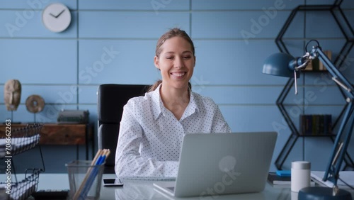 young woman in white shirt sits at table in office with laptop, typing on keyboard concentrated. smiles and rejoices to success result and great news, looking to monitor