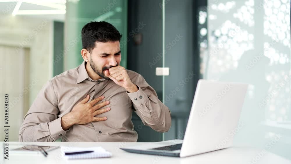 Handsome businessman in a shirt coughing while sitting at workplace at desk in business office. Sick with a cold or flu employee feels bad, holds his hand to his chest. Bearded male has a bronchitis Handsome businessman in a shirt coughing while sitting at workplace at desk in business office. Sick with a cold or flu employee feels bad, holds his hand to his chest. Bearded male has a bronchitis