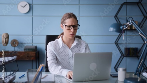 young woman in white shirt and eyeglasses sit at table desk in office, working at laptop. frustrated and worried, frowns, gives up and closes computer to have a break and breath out. photo