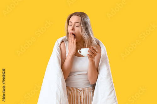 Sleepy young woman with blanket and cup of coffee on yellow background