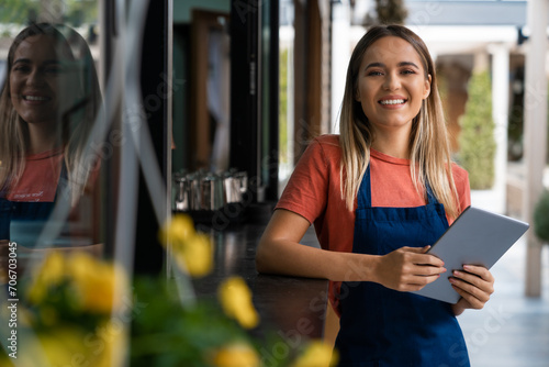 Smiling confident young woman wearing apron looking at camera holding digital tablet fintech device standing outside the restaurant or Cafe.