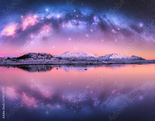 Milky Way arch over snowy mountains and sea coast at winter night. Landscape with snowy rocks, pink starry sky, reflection in water, fjord. Lofoten Islands, Norway. Space. Beautiful arched milky way