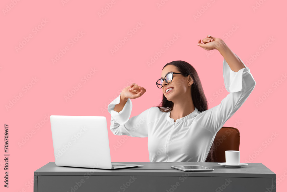 Tired young female programmer stretching at table with laptop on pink background Tired young female programmer stretching at table with laptop on pink background